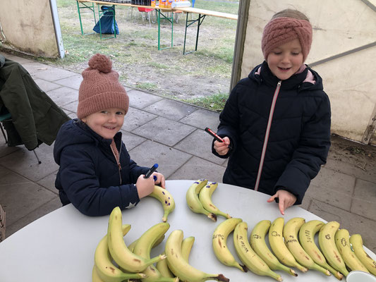 Zwei Kinder in Winterkleidung stehen an einem runden Tisch und bemalen mit Filzstiften Bananen, die in einer Reihe vor ihnen liegen.