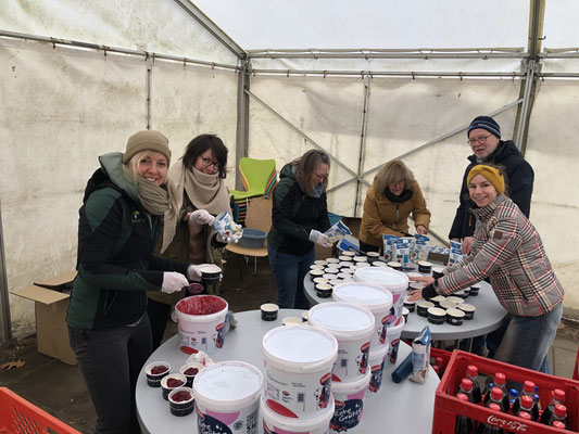 Sechs Erwachsene stehen in einem Zelt um Tische und befüllen kleine Becher mit Milchreis und Roter Grütze aus großen Eimern und Tetrapacks. Sie tragen warme Kleidung und Einweghandschuhe.