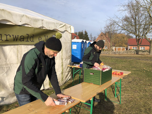 Zwei Männer in grünen Jacken stehen vor einem Zelt mit dem Banner "Naturwald e.V." und bereiten auf Holztischen im Freien eine Verpflegungsstation mit Obst und Müsliriegeln vor. Im Hintergrund stehen blaue mobile Toilettenkabinen und Häuser.