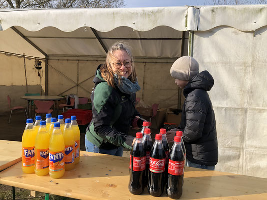 Eine Frau und ein Junge richten draußen vor einem Zelt, in dem ein Tisch mit Stühlen steht, eine Getränkestation mit Fanta- und Cola-Flaschen auf einem Holztisch ein. Die Frau schaut in die Kamera und lächelt.