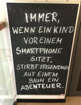 Schwarze Kreidetafel mit weißer handschriftlicher Aufschrift in großen Druckbuchstaben: „Immer, wenn ein Kind vor einem Smartphone sitzt, stirbt irgendwo auf einem Baum ein Abenteuer.“ Die Tafel lehnt schräg an einer Wand in einem Innenraum.