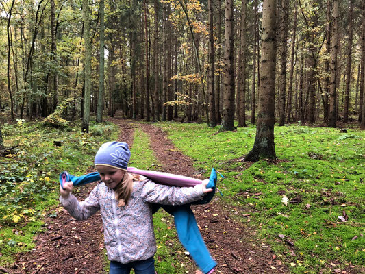 Ein Mädchen in einer hellen Jacke und mit einer Mütze läuft einen bewaldeten Weg entlang und trägt eine blaue Jacke über den Schultern.