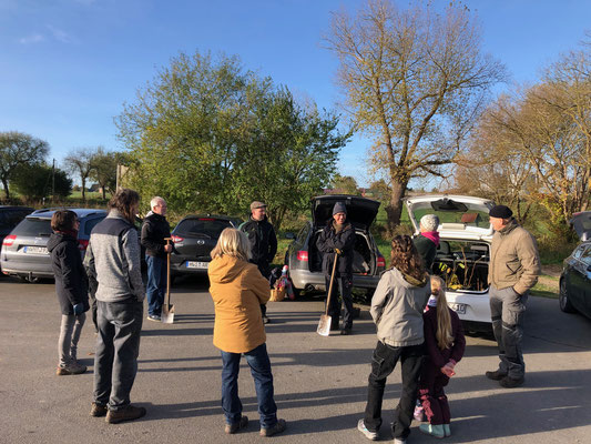 Eine Gruppe von Menschen mit Schaufeln steht auf einem Parkplatz in einem Kreis versammelt. Im Hintergrund stehen Autos mit geöffneten Kofferräumen.